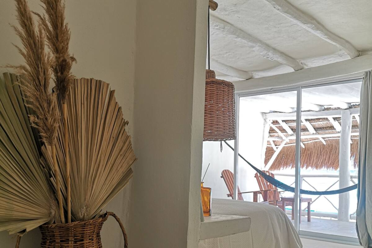 Una habitación con una cesta de mimbre con plumas de pluma de pampa, una lámpara colgante de mimbre y vistas a una zona de estar exterior con techo de paja.