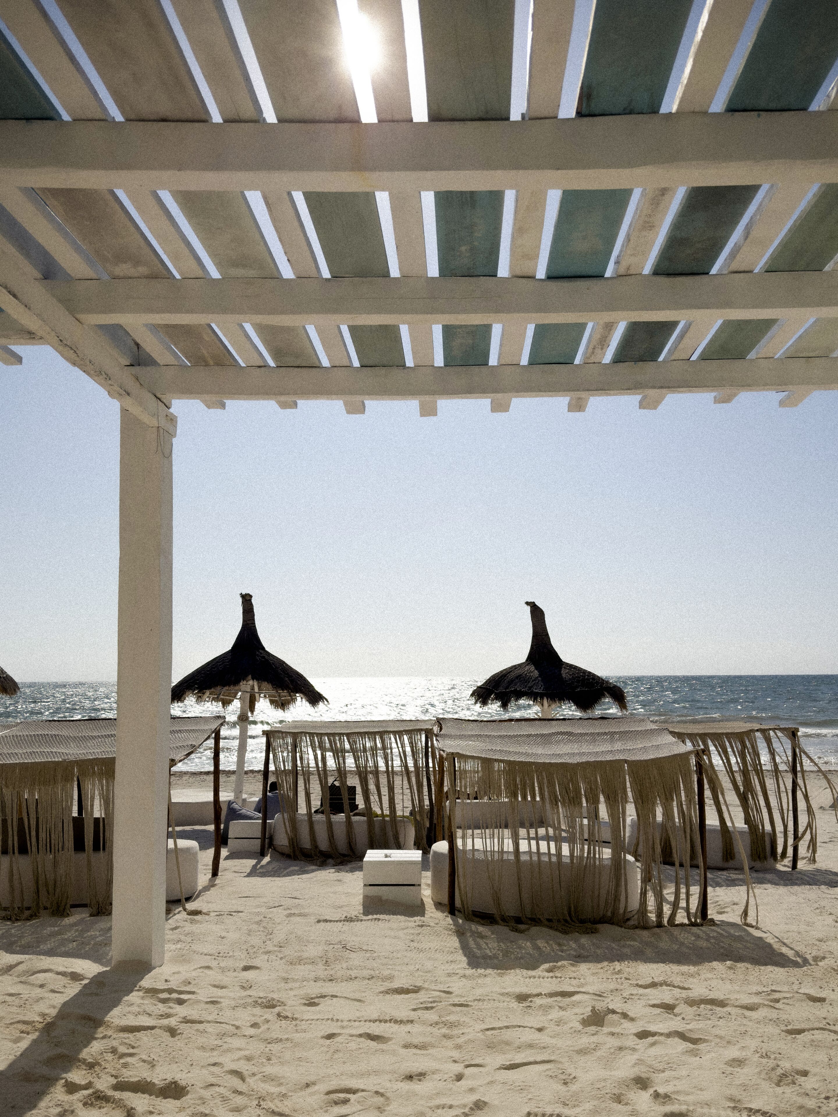 A beach with thatched umbrellas and lounge chairs under a pergola.