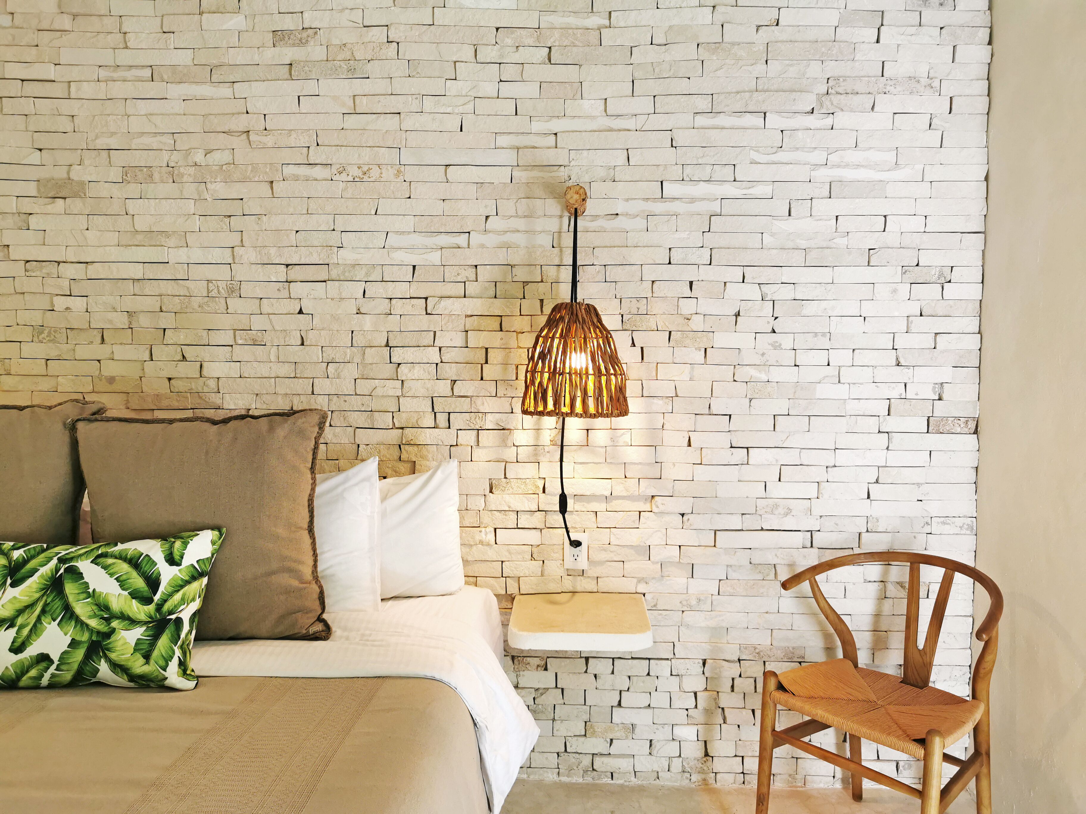 Quarto com parede em tijolos, cama com roupa de cama branca e marrom, cadeira de madeira e luminária pendente.