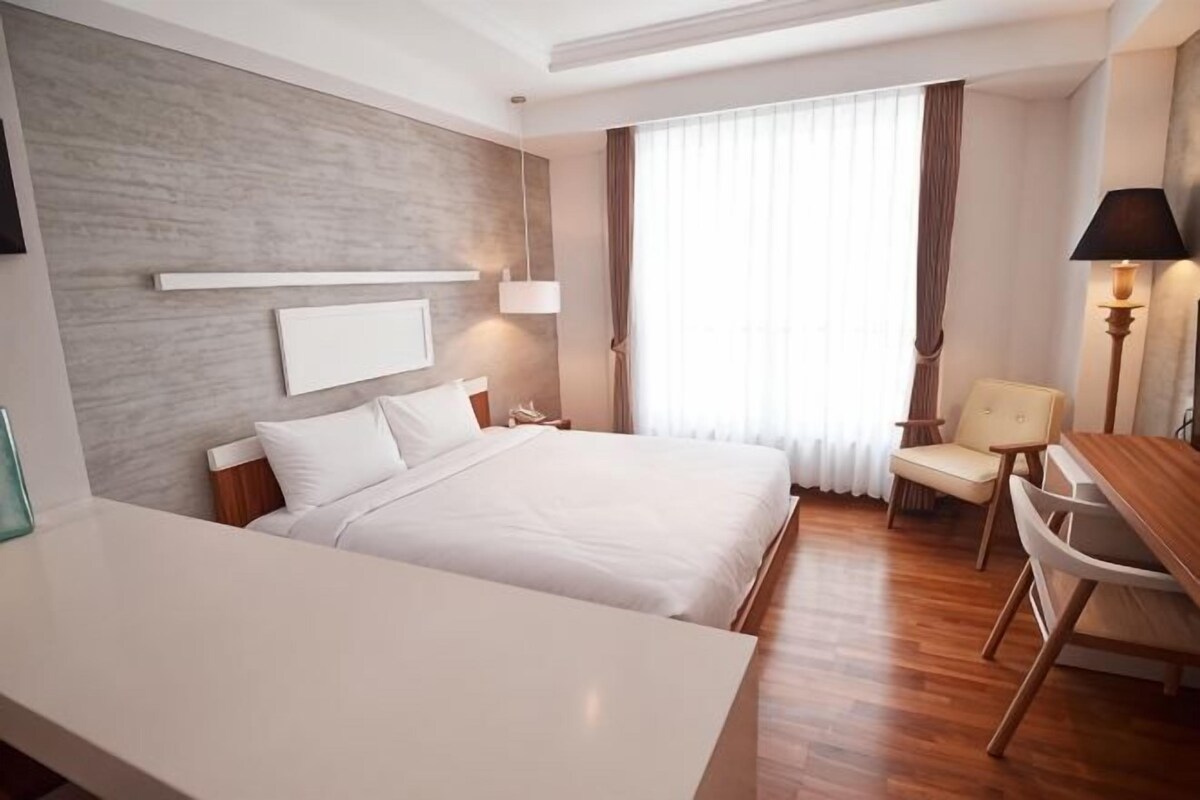A hotel room with a bed, bedside table, chair, desk, lamp, and a window with curtains.