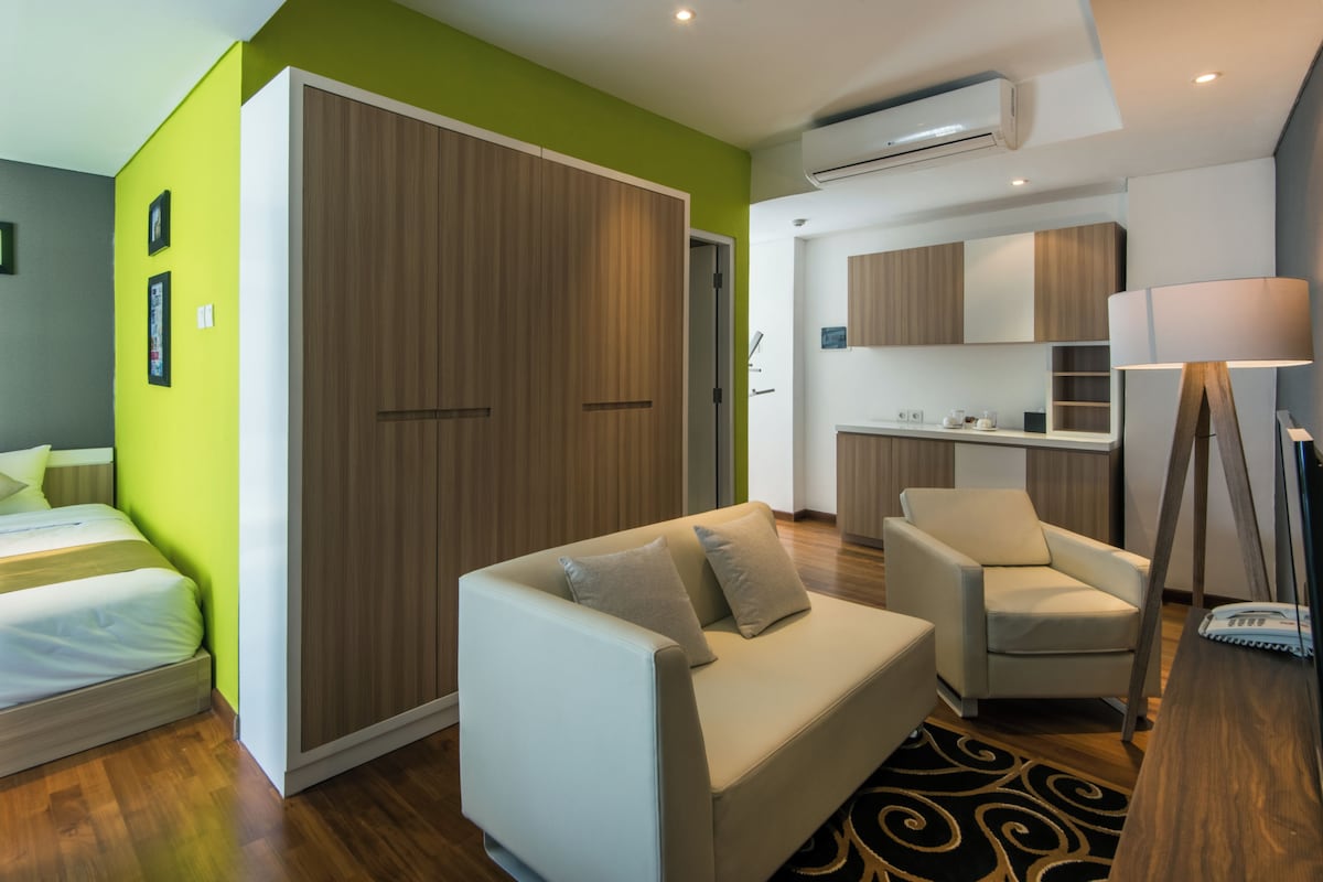 A modern hotel room with a bed, a sofa, a chair, a kitchenette, and a large wardrobe.