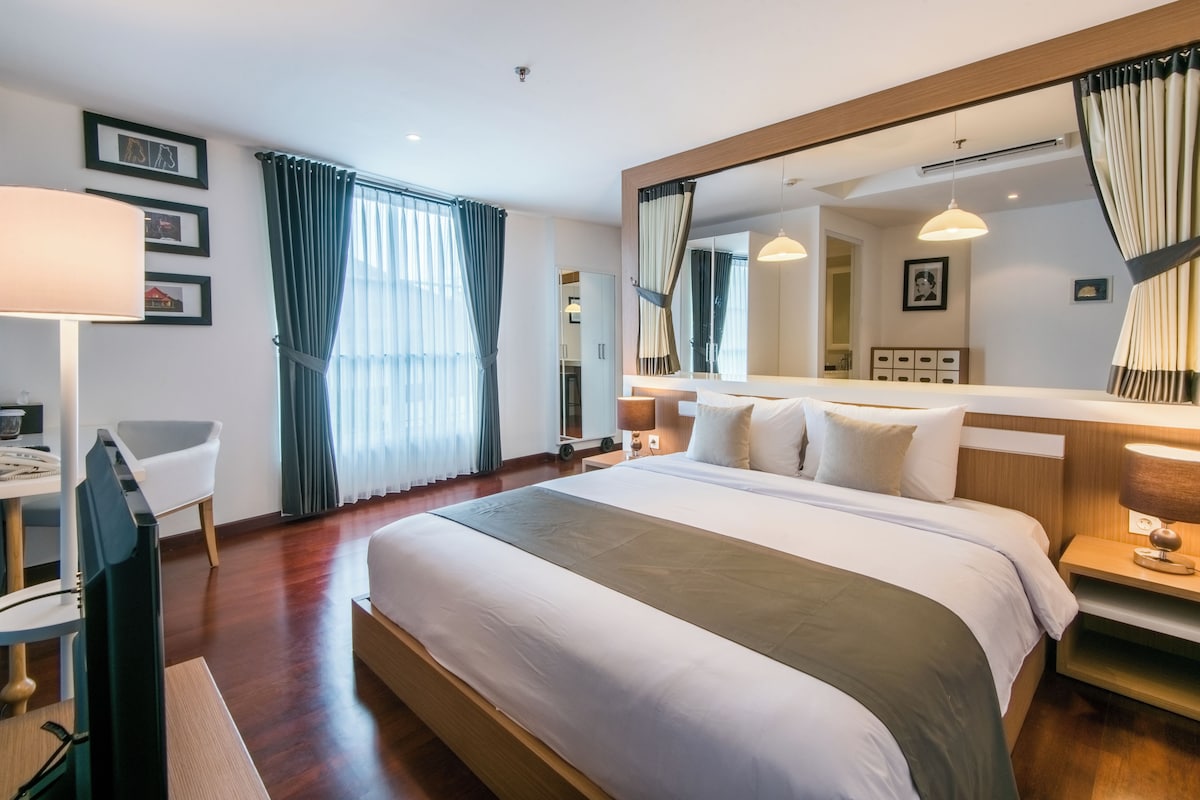 A hotel room with a large bed, a desk with a chair, a mirror, and a window with curtains.