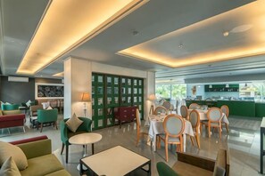 A modern hotel lobby with a dining area, seating arrangements, and a green wall with framed pictures. - Yan's House Hotel (Kuta)