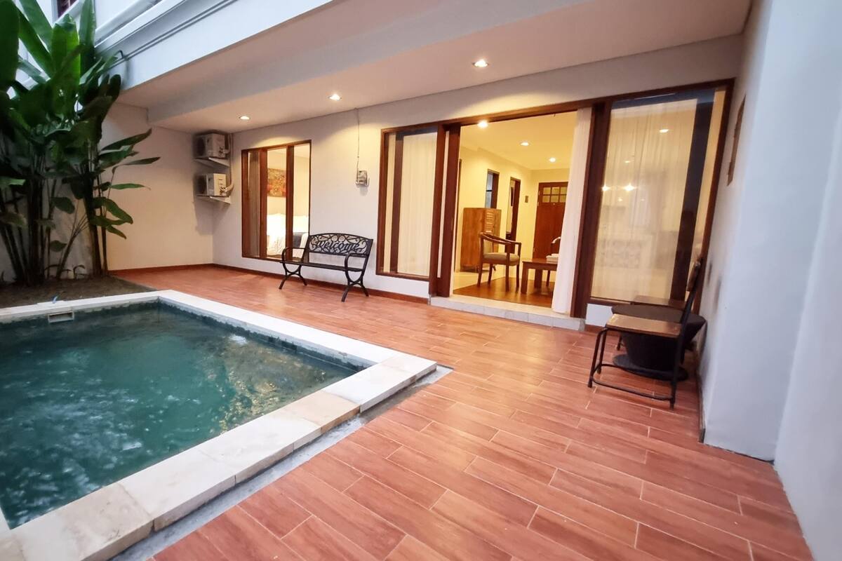 Ein Schwimmbecken mit gefliestem Deck, einer Bank und einer Tür zu einem Innenbereich mit Stuhl und Tisch.