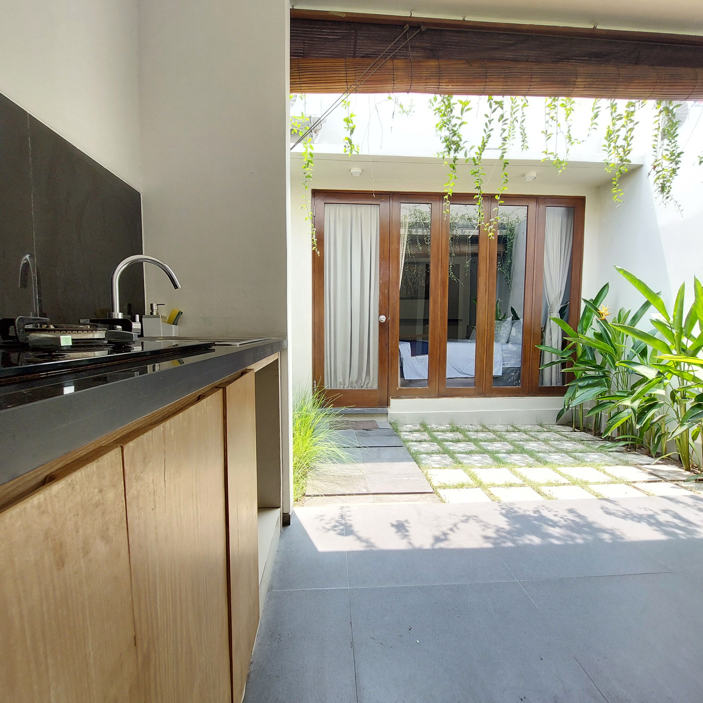 A modern kitchen with a built-in stove and sink, a wooden cabinet, and a sliding glass door leading to a patio with a tiled floor and potted plants.
