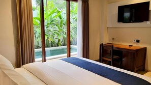 A hotel room with a bed, a desk, a TV, and a view of greenery and a pool. - Omatha Village (Denpasar)