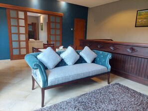A blue sofa with patterned cushions, a wooden bench, and a patterned rug in a room with a tiled floor and a door leading to another room.