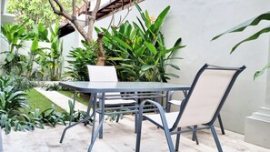 A patio with a metal table and chairs, surrounded by lush greenery and a white wall.