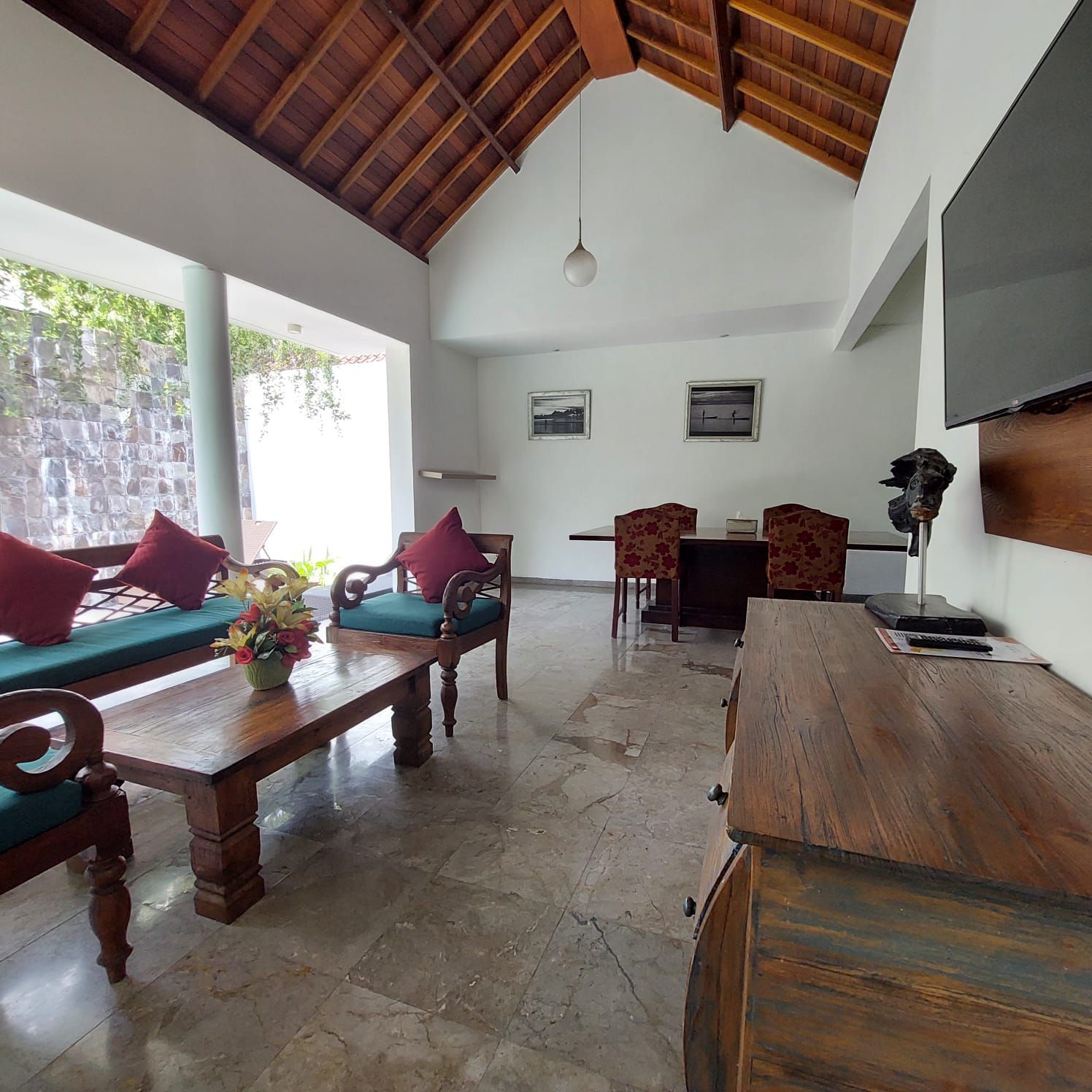 A spacious room with a high ceiling, a wooden dining table, two sofas with cushions, a wooden cabinet, and a television mounted on the wall.