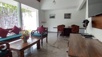 A spacious room with a high ceiling, a wooden dining table, two sofas with cushions, a wooden cabinet, and a television mounted on the wall.