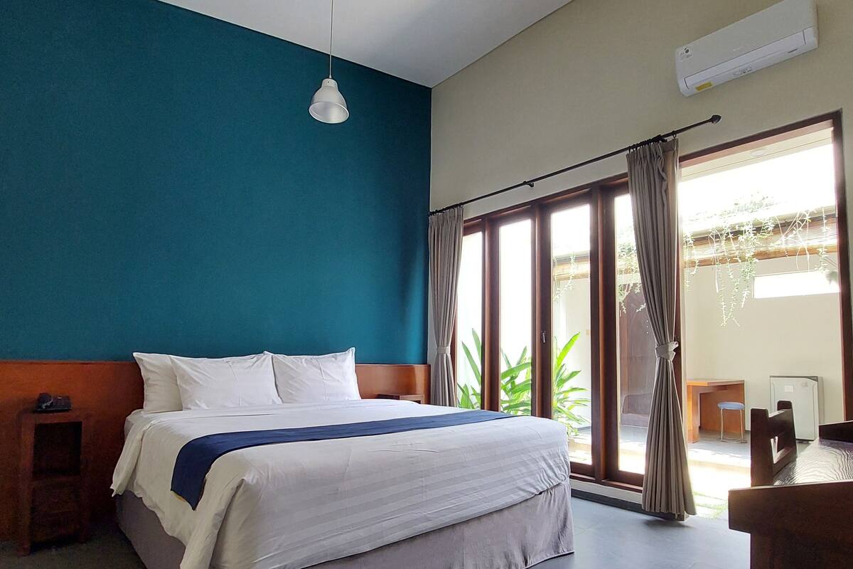 A hotel room with a bed, a desk, and a chair. There is a window with curtains and a view of greenery outside.