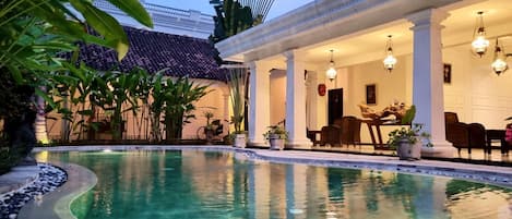 A pool with a tiled deck, a covered seating area with chairs and tables, and a building with a tiled roof.