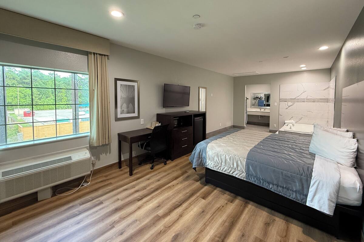Quarto de hotel com cama, escrivaninha, cadeira, televisão e banheiro com espelho e pia.