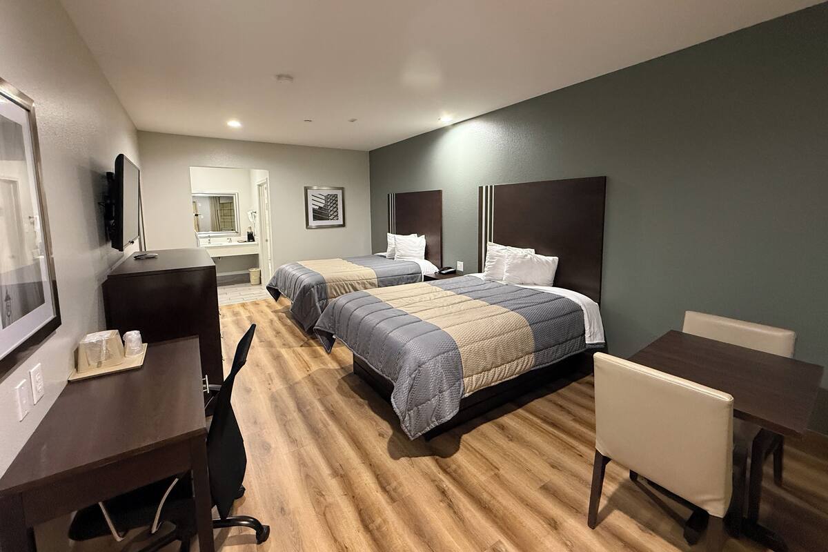 Quarto de hotel com duas camas, uma escrivaninha, uma cadeira, uma televisão e um banheiro visível através de uma porta aberta.