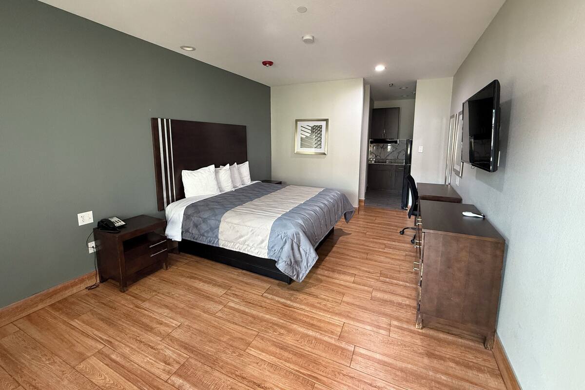 Quarto de hotel moderno com uma cama grande, uma escrivaninha, uma cadeira, televisão de tela plana e uma kitchenette ao fundo.