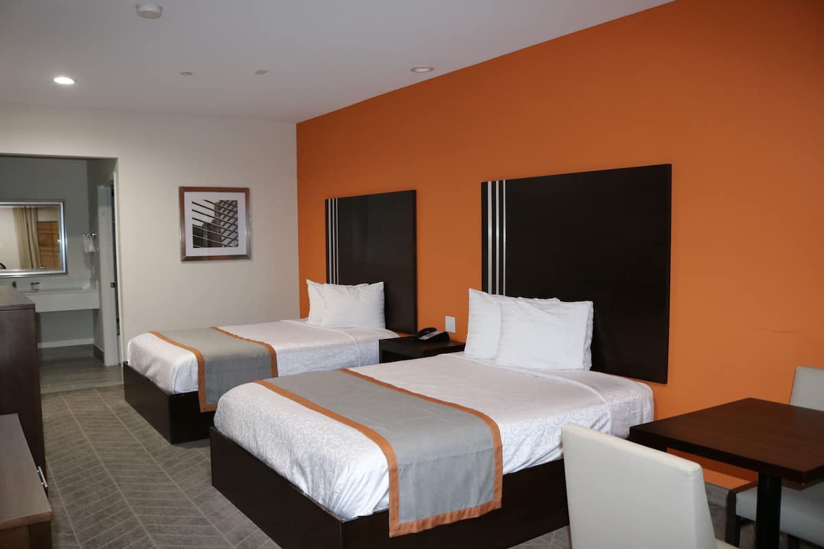 Quarto de hotel com duas camas, uma escrivaninha, uma cadeira, uma mesinha de cabeceira, um espelho e uma parede com detalhes em laranja.
