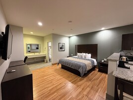 Una habitación de hotel moderna con suelo de madera, una cama grande con cabecera, un escritorio con silla, un televisor y un baño con lavabo e inodoro.