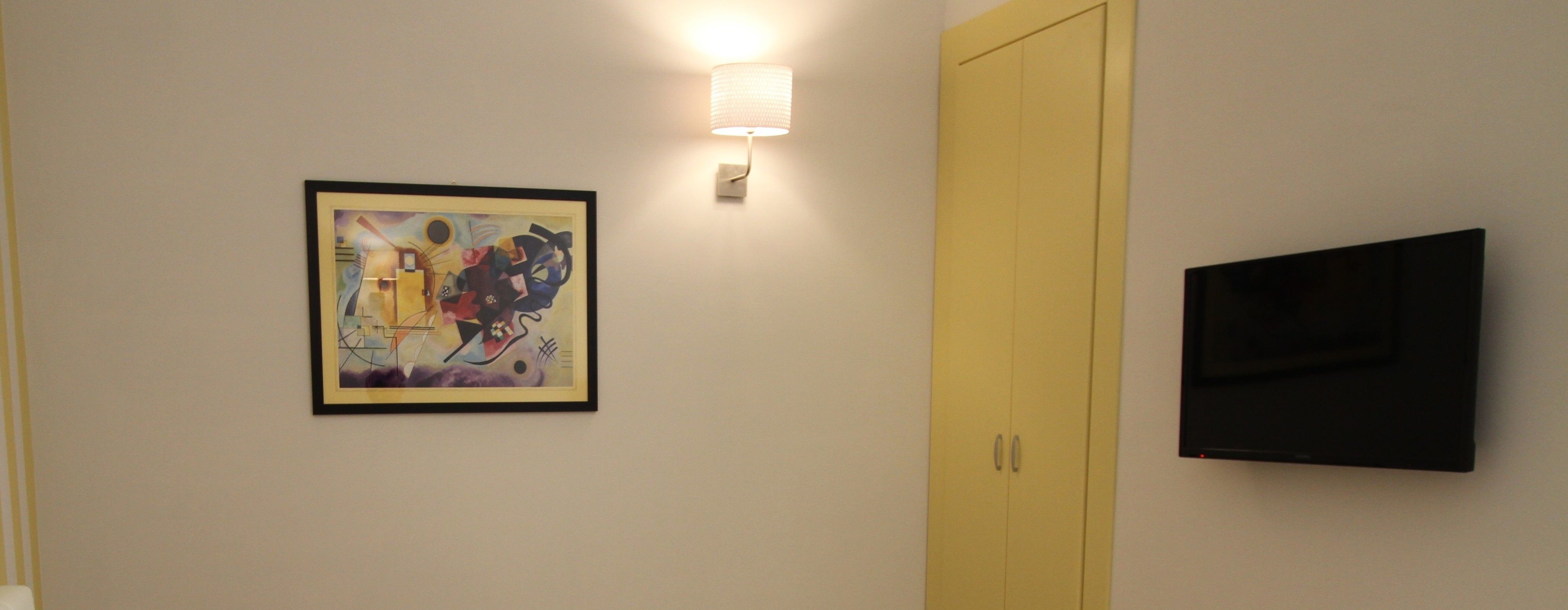 Una habitación con un cuadro abstracto enmarcado, una lámpara de pared, una puerta amarilla cerrada y un televisor de pantalla plana.