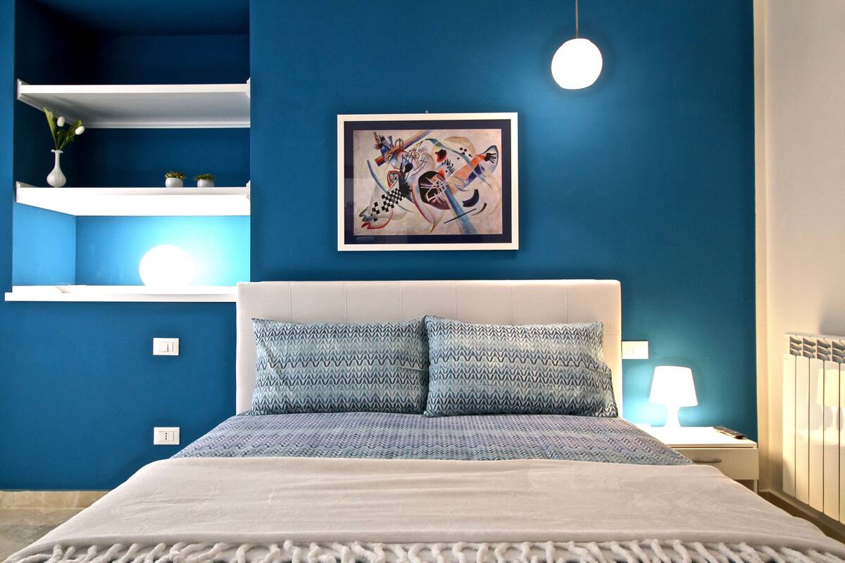 Un dormitorio moderno con una pared azul de acento, un cabecero blanco, una colcha estampada y un cuadro abstracto enmarcado sobre la cama.