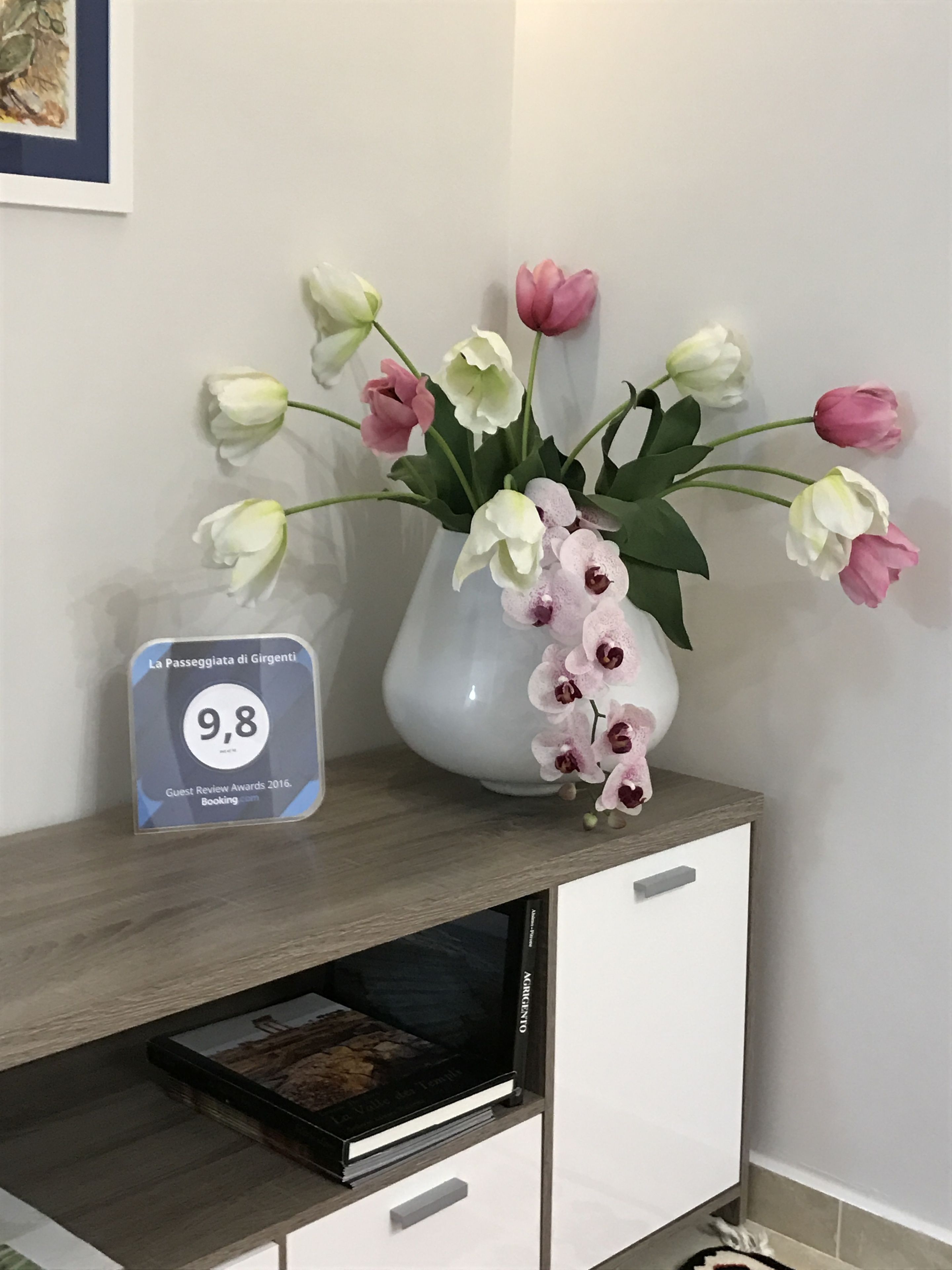 Un jarrón blanco con flores rosadas y blancas sobre una repisa de madera, un certificado enmarcado y un libro.