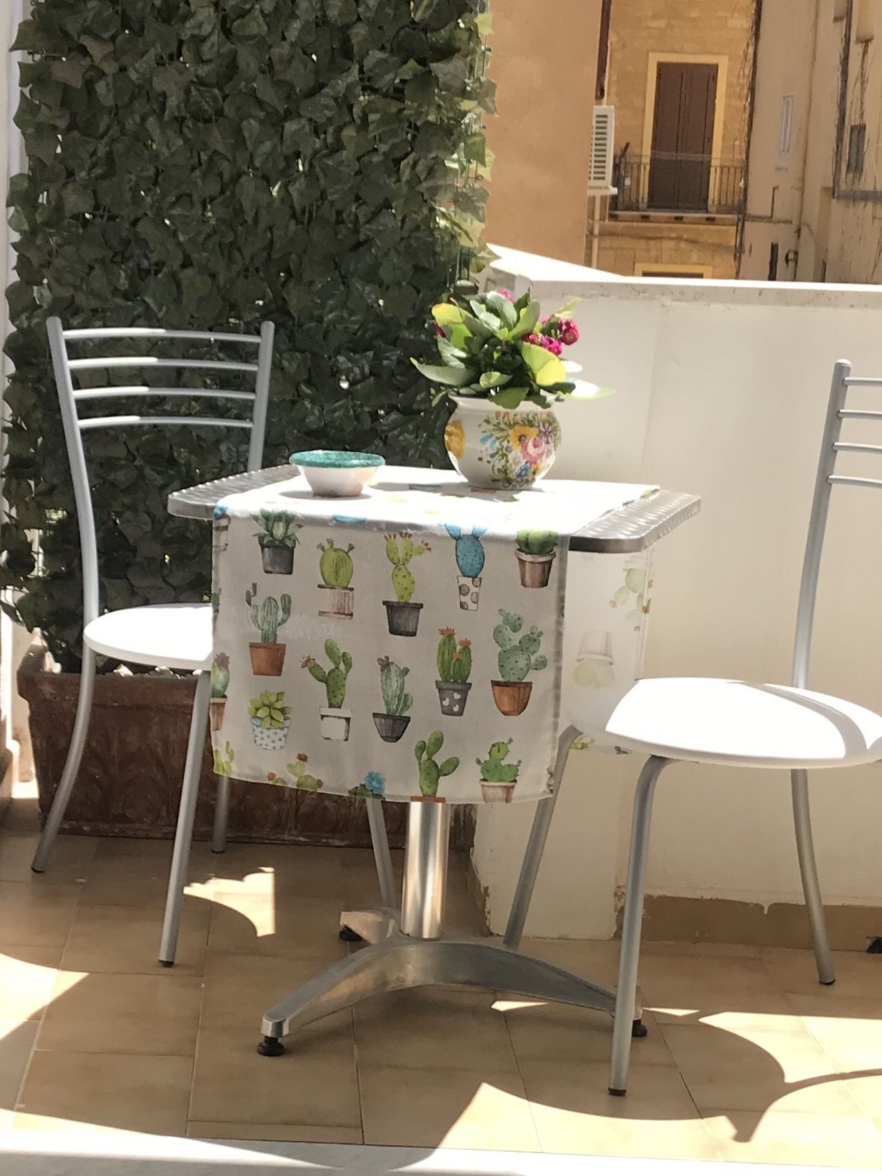 Ein kleiner Tisch im Freien mit Tischdecke im Kaktus-Design, zwei weiße Stühle und ein Topfpflanze.