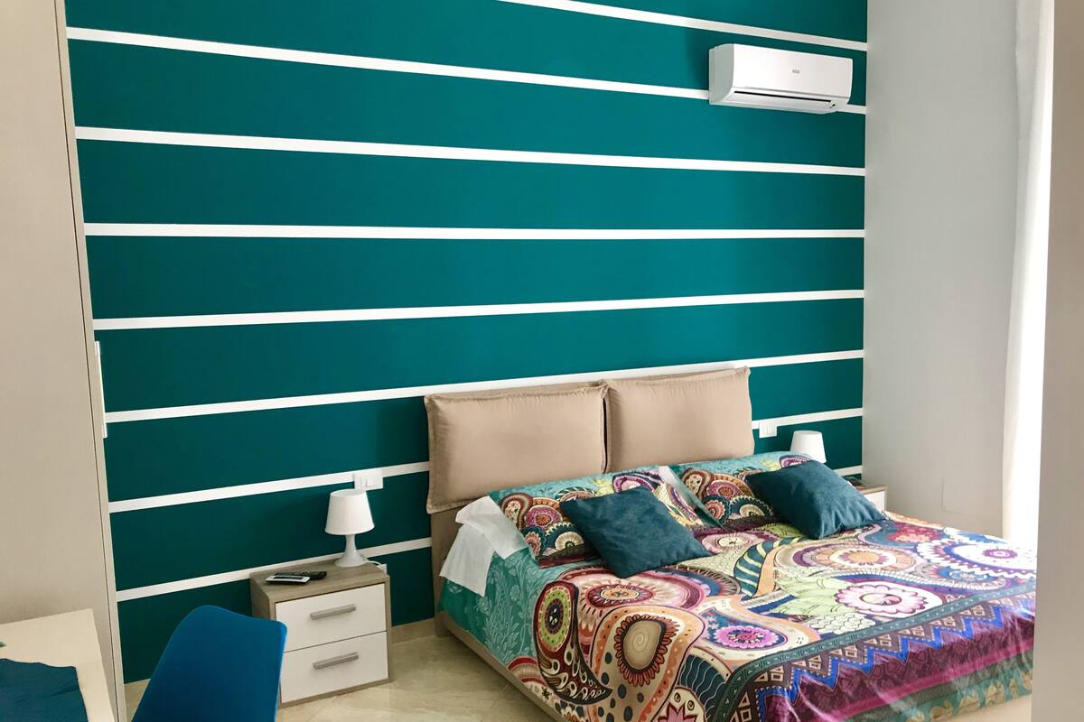 Un dormitorio con una pared color turquesa, una cama con edredón estampado y colorido, una mesita de noche con lámpara y un mueble auxiliar con cajones.
