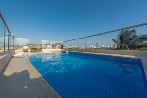 Una piscina en la azotea con agua azul cristalina, rodeada de barandillas de vidrio, que ofrece vistas del horizonte de la ciudad.