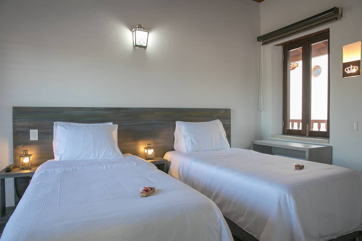 Deux lits simples avec du linge de lit blanc, des tables de chevet avec des lampes, une tête de lit en bois et une fenêtre donnant sur l’extérieur.