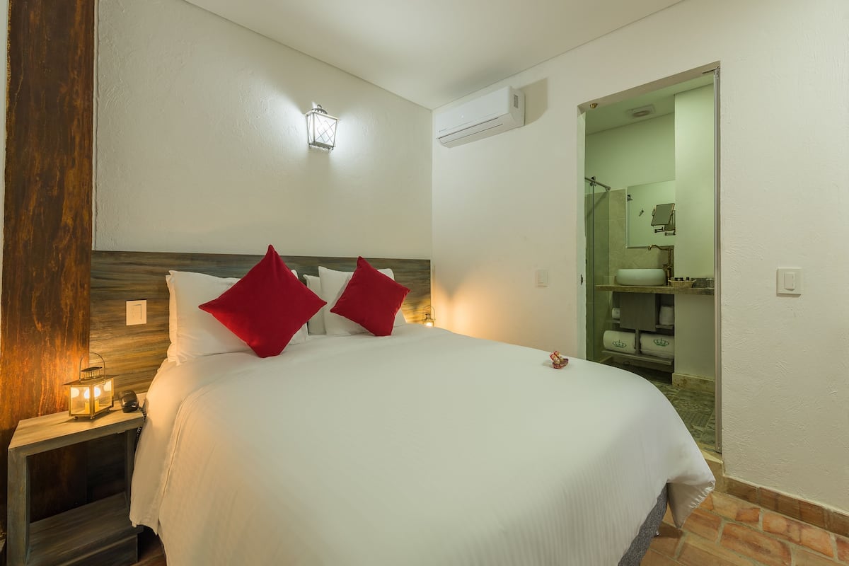 Une chambre d’hôtel avec un lit, des oreillers rouges, une table de chevet avec une lampe, un luminaire fixé au mur et un appareil de climatisation.