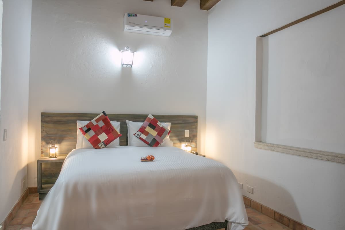 Un lit bien fait, recouvert d’une couvre-lit blanche et agrémenté de deux coussins décoratifs, une table de chevet avec une lampe, et un climatiseur fixé au mur.