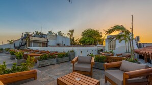 Une terrasse sur le toit, aménagée avec des meubles d’extérieur, notamment des chaises et des tables en bois, entourée de plantes en pot et offrant une vue sur le paysage urbain.