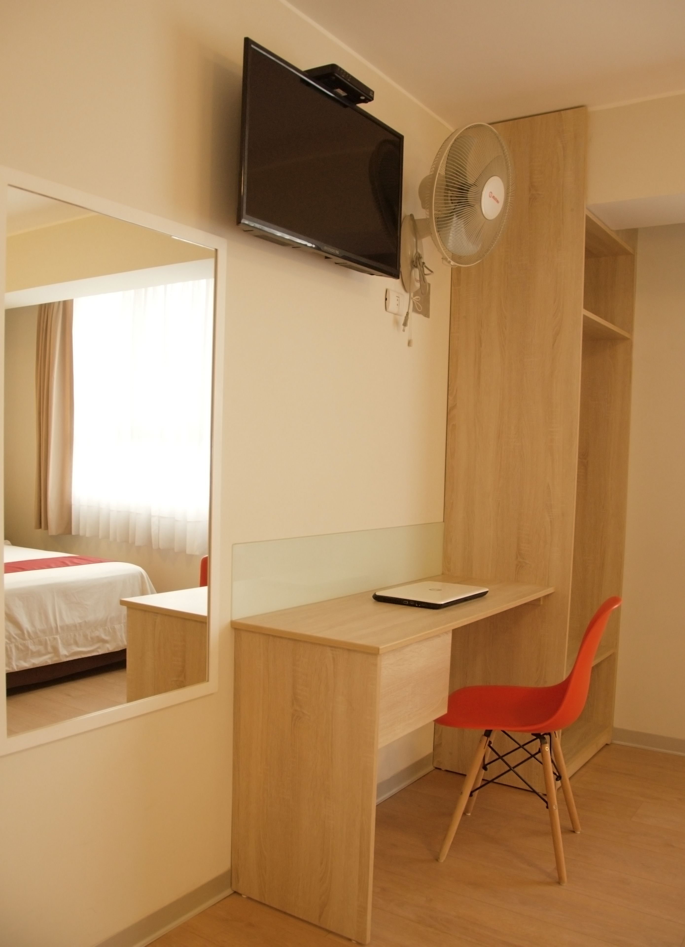 A hotel room with a wooden desk, a red chair, a TV mounted on the wall, and a large window with curtains.