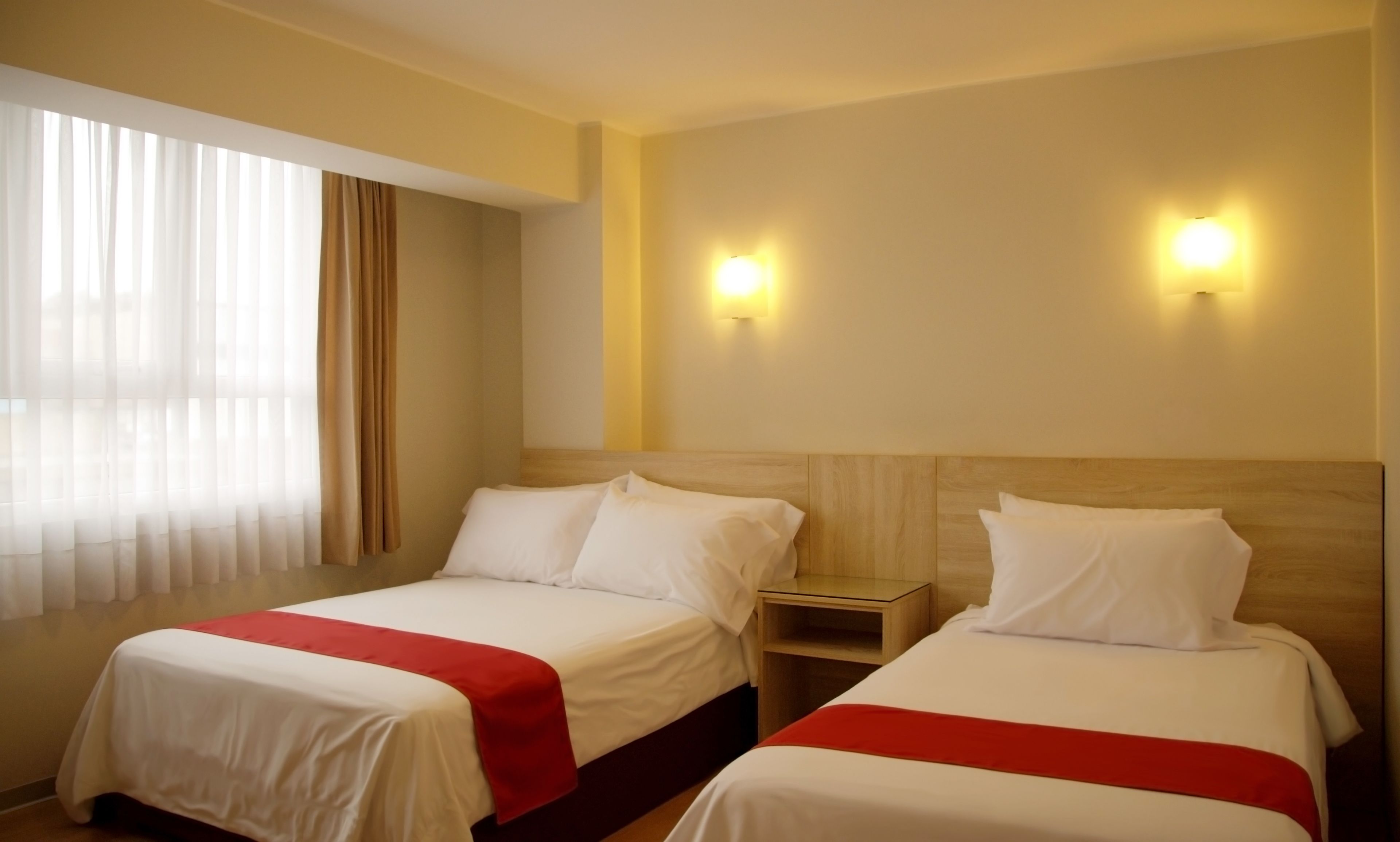 Ein Hotelzimmer mit zwei Betten, jeweils mit Holz Kopfteil und weißer Bettwäsche. Zwei Wandleuchten und ein Fenster mit durchscheinenden Vorhängen.