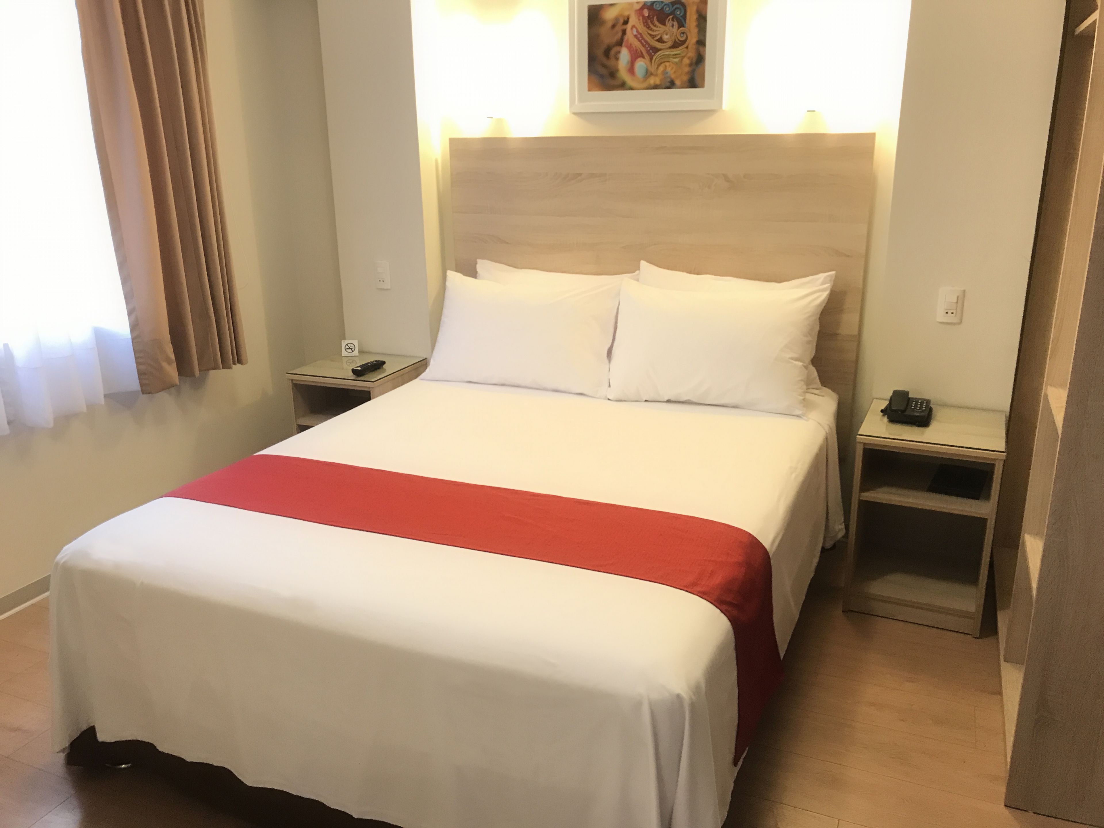A hotel room with a large bed, a red accent on the bedspread, two bedside tables with lamps, and a painting on the wall.
