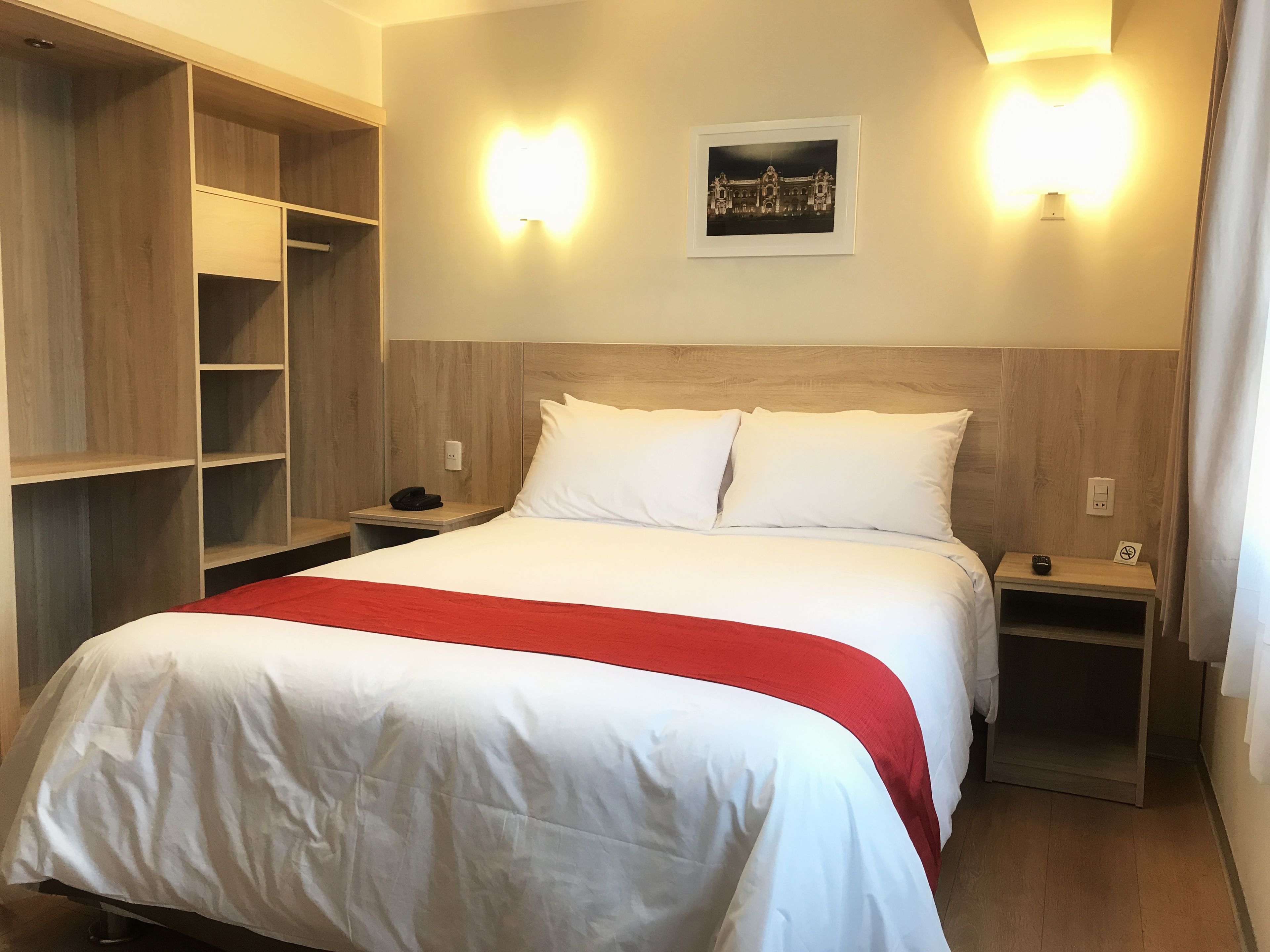 A hotel room with a large bed, wooden headboard, two bedside tables, a nightstand, a wardrobe, and two wall-mounted lights.