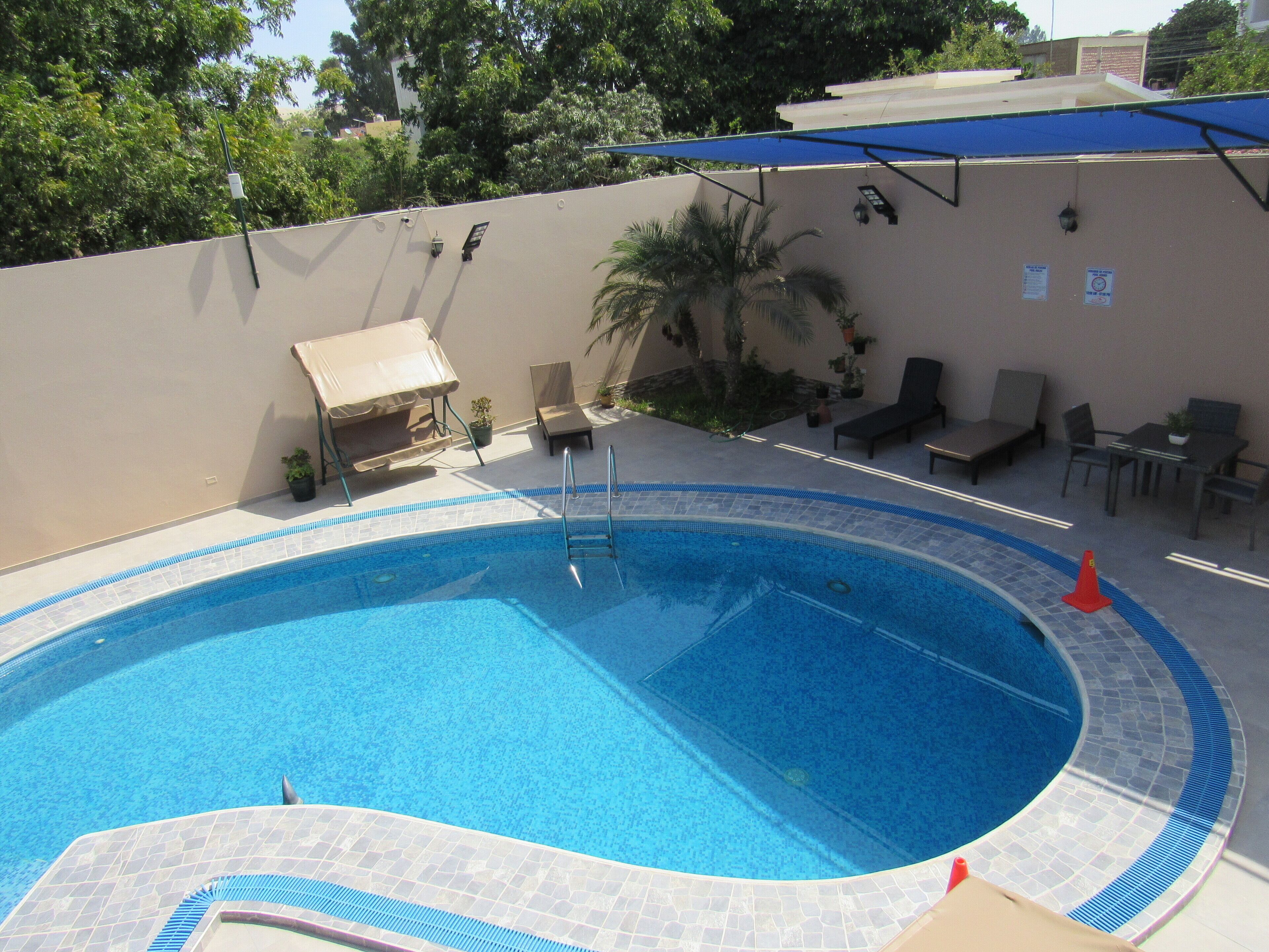 A swimming pool with a tiled edge, a lounge area with chairs and a table, and a covered seating area.