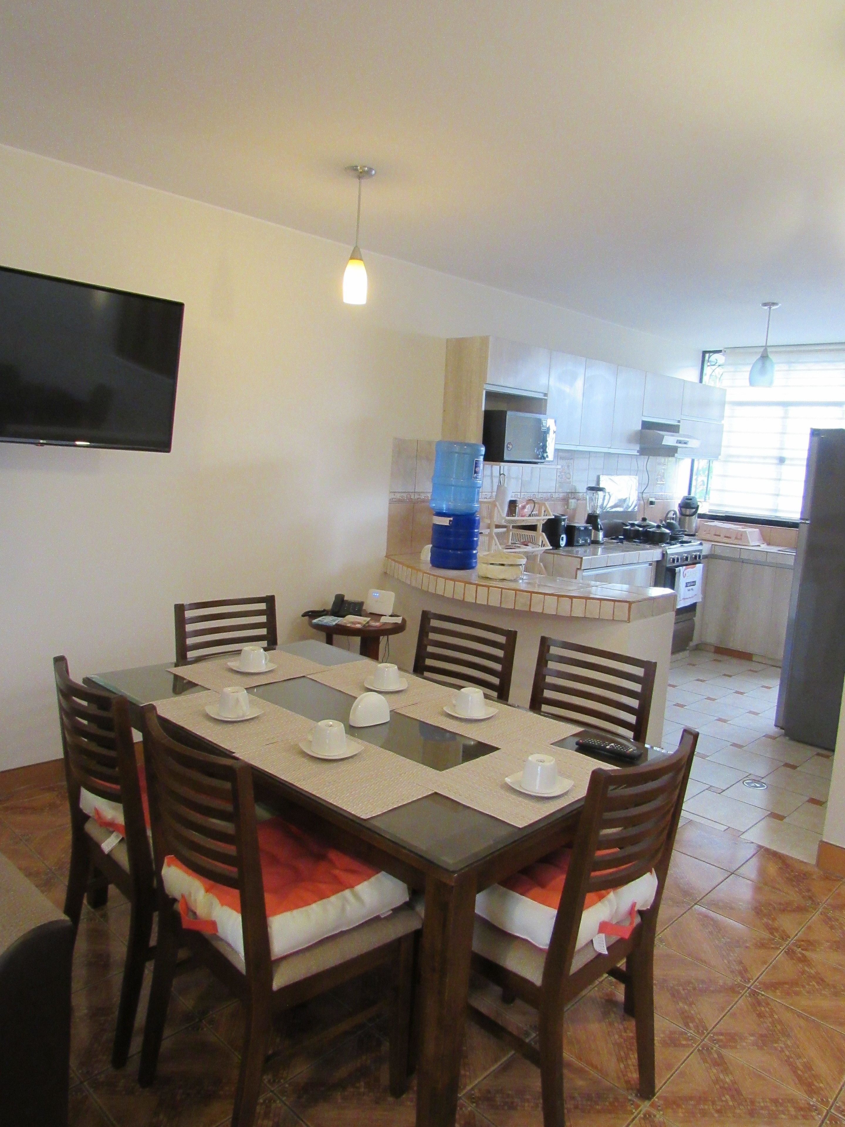 Comedor con una mesa dispuesta para cuatro personas, una cocina al fondo y un televisor montado en la pared.