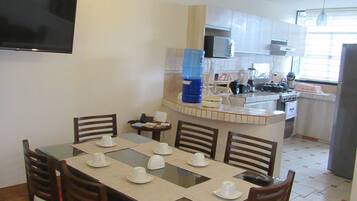 Comedor con una mesa dispuesta para cuatro personas, una cocina al fondo y un televisor montado en la pared.