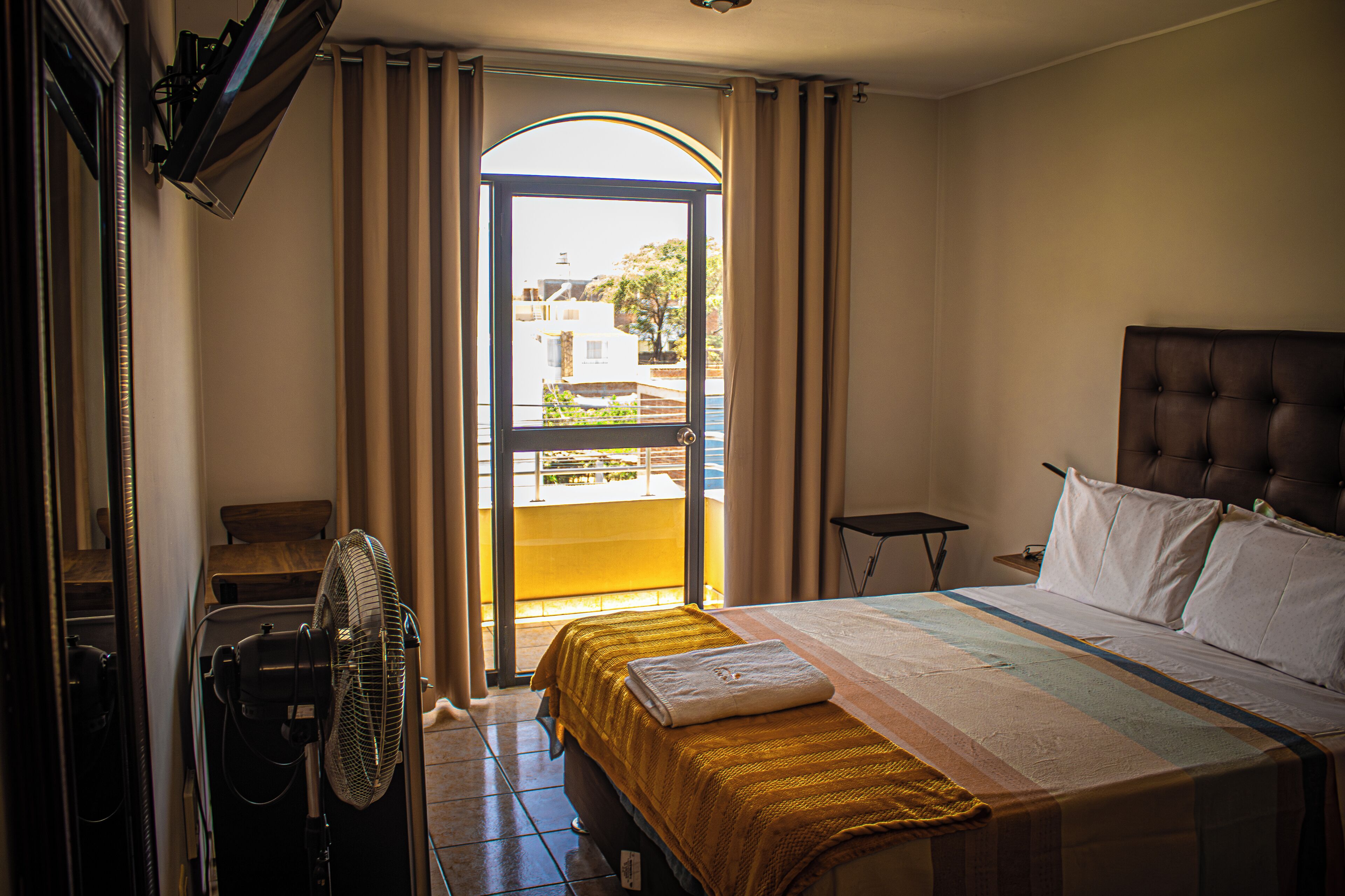 A hotel room with a bed, a fan, a window with curtains, and a view of buildings outside.