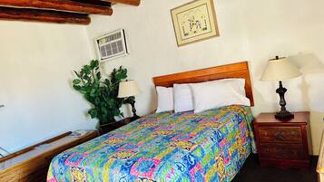 Um quarto com uma cama de madeira, um edredom colorido, uma mesa de cabeceira com uma luminária, uma planta em vaso e um ar condicionado fixado na parede.