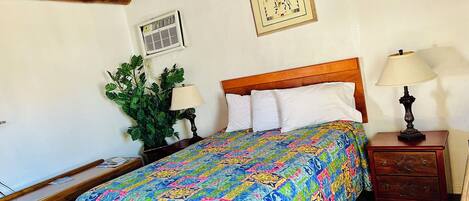 Un dormitorio con una cama de madera, un edredón colorido, una mesita de noche con una lámpara, una planta en maceta y un aire acondicionado instalado en la pared.