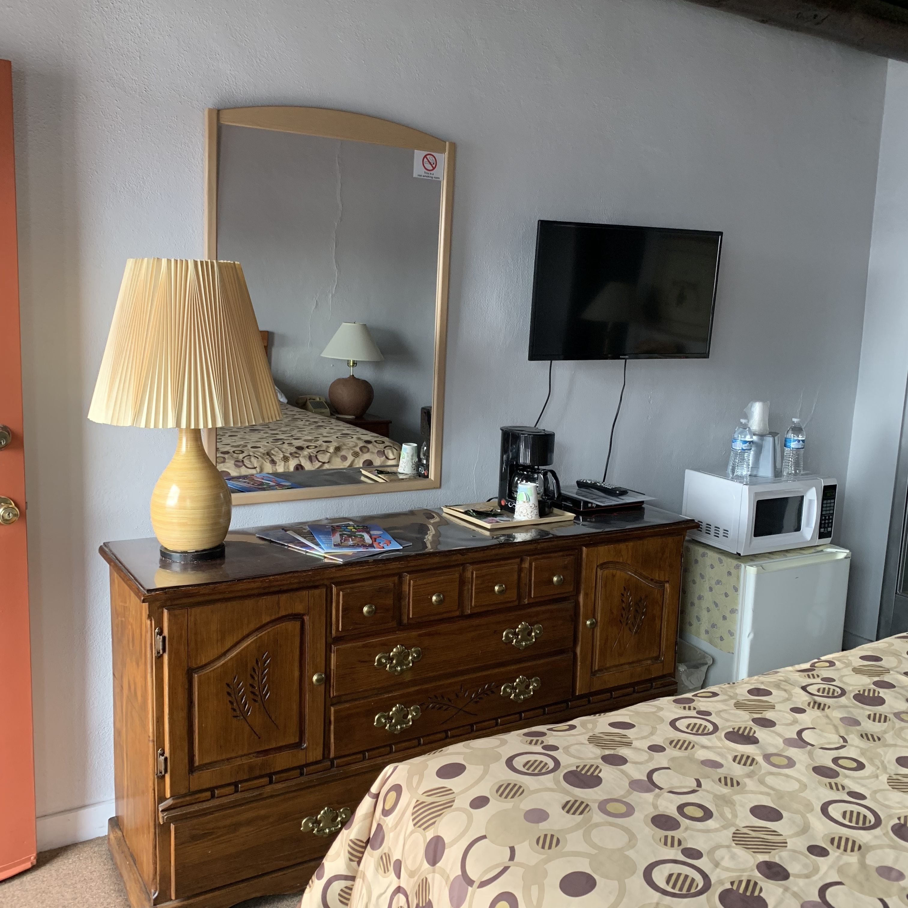 Ein Hotelzimmer mit einem hölzernen Nachttisch, einem Bett mit gemusterten Bettbezügen, einem an der Wand befestigten Fernseher und einem Kühlschrank.