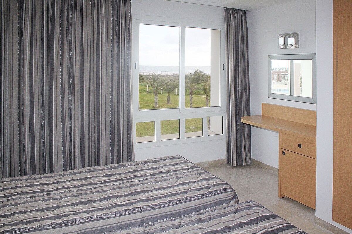 A bedroom with a bed, a desk, and a window with a view of palm trees and a building.
