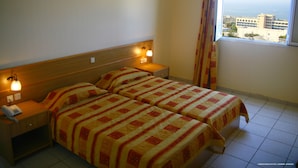 A hotel room with two beds, a wooden headboard, bedside lamps, a telephone on the nightstand, and a window with a view of buildings and the sea.