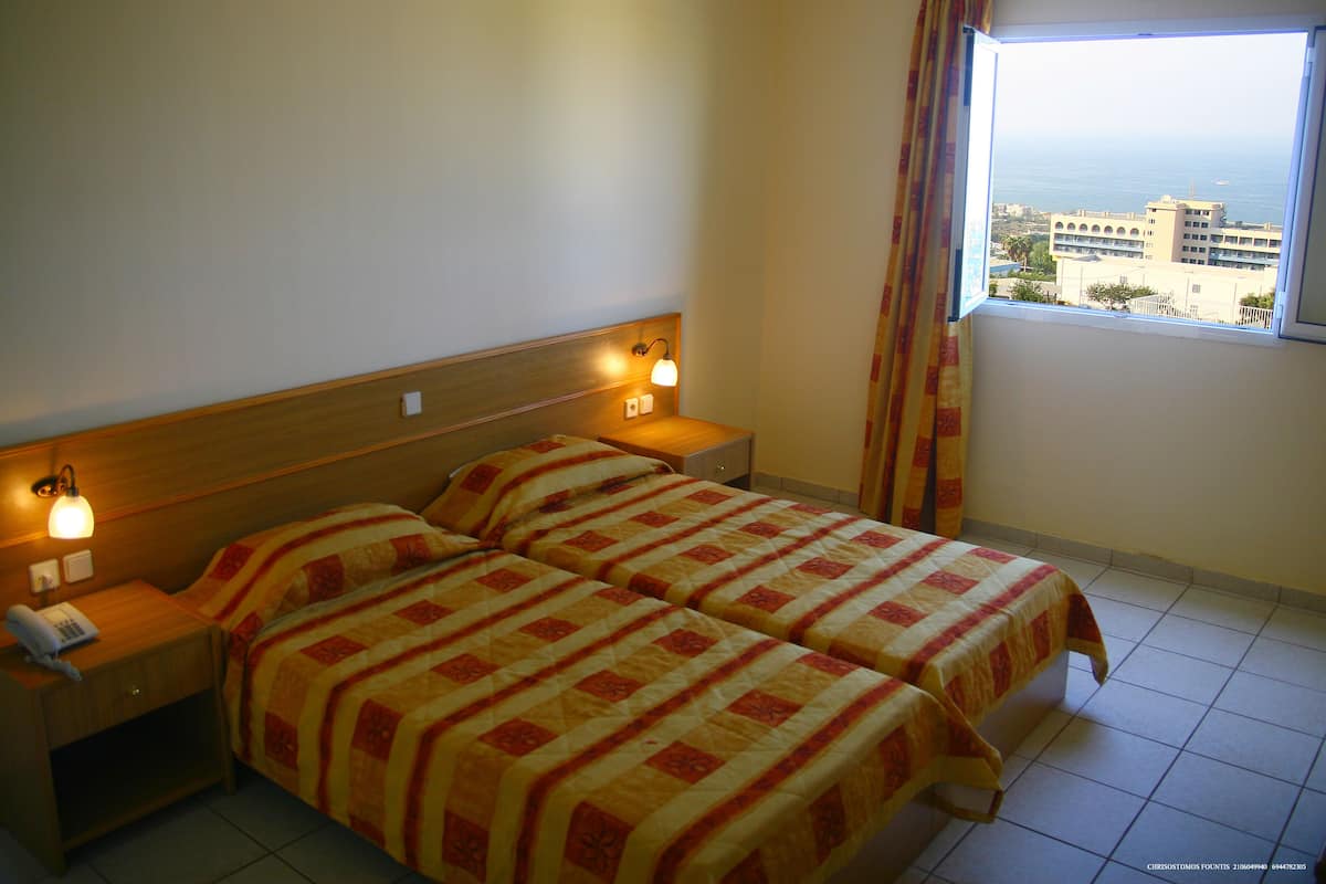 A hotel room with two beds, a wooden headboard, bedside lamps, a telephone on the nightstand, and a window with a view of buildings and the sea.