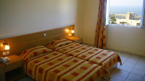 A hotel room with two beds, a wooden headboard, bedside lamps, a telephone on the nightstand, and a window with a view of buildings and the sea. - Aqua Sun Village (Hersonissos)