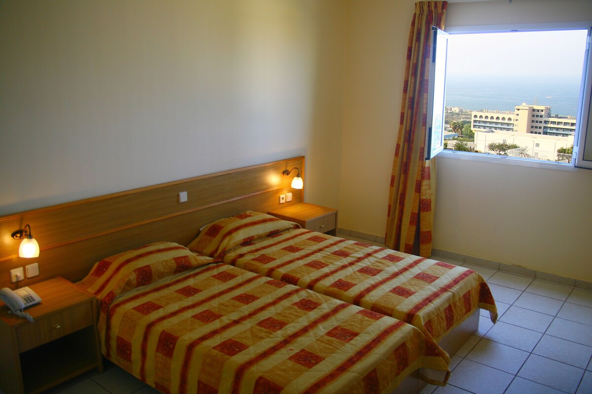 A hotel room with two beds, a wooden headboard, bedside lamps, a telephone on the nightstand, and a window with a view of buildings and the sea.