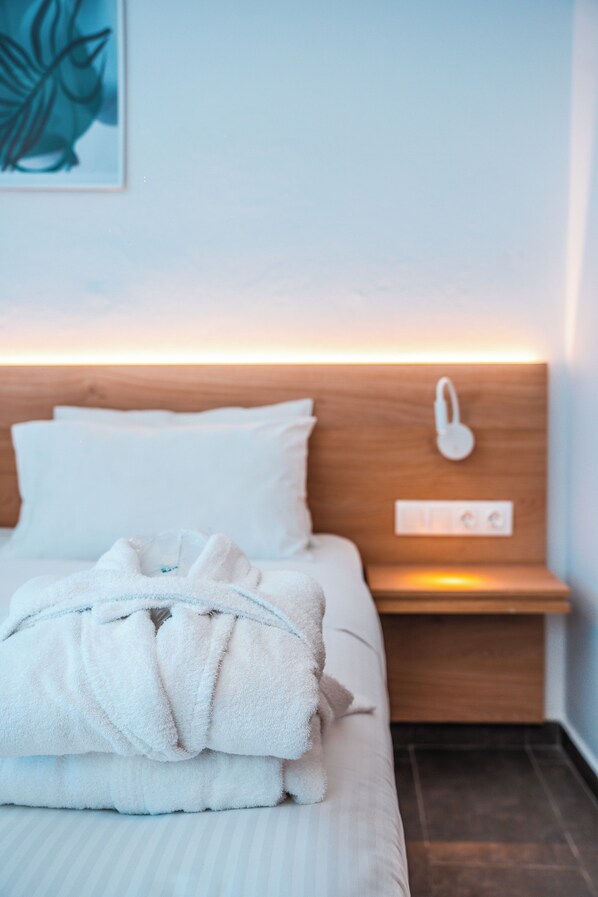 A neatly made bed with a white duvet and a white bathrobe, a bedside table with a lamp, and a wall-mounted light switch. - Kahlua Boutique Hotel (Hersonissos)