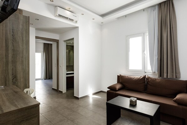 A modern living room with a brown sofa, a coffee table, and a large window with curtains. There is a hallway leading to another room and an air conditioning unit on the wall.