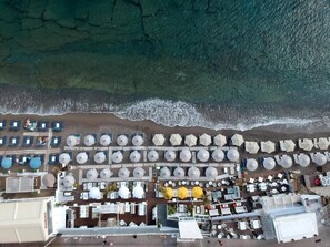 Vue aérienne d’un complexe hôtelier situé en bord de mer, avec des rangées de parasols et de chaises longues, un espace de restauration et un bâtiment doté d’un toit plat.